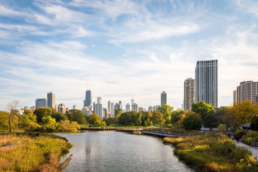 Chicago city skyline with tall buildings in the background, trees and greenery along a river in the foreground under a blue sky with wispy clouds.