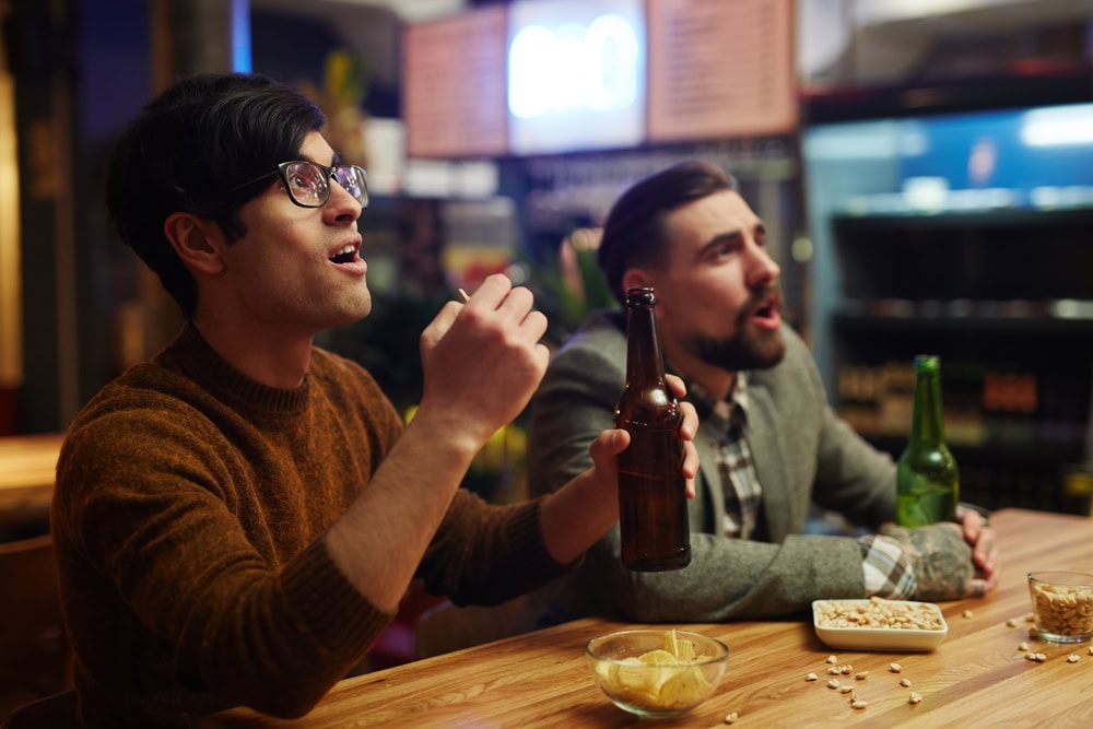 Two men sit at a bar watching something off-camera, one holding a beer and the other eating snacks; snack bowls and bottles are on the wooden counter.