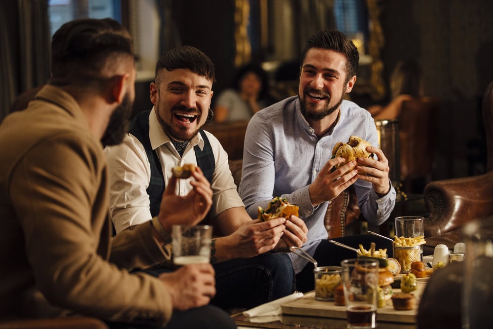 Three men sit at a table in a restaurant, eating burgers and fries, smiling and talking. Glasses of drinks are on the table.