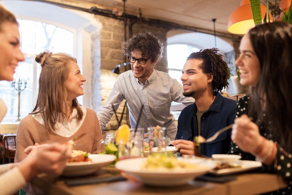Five people sitting at a restaurant table, eating and smiling, with one person standing and engaging with the group. Plates of food and drinks are on the table.