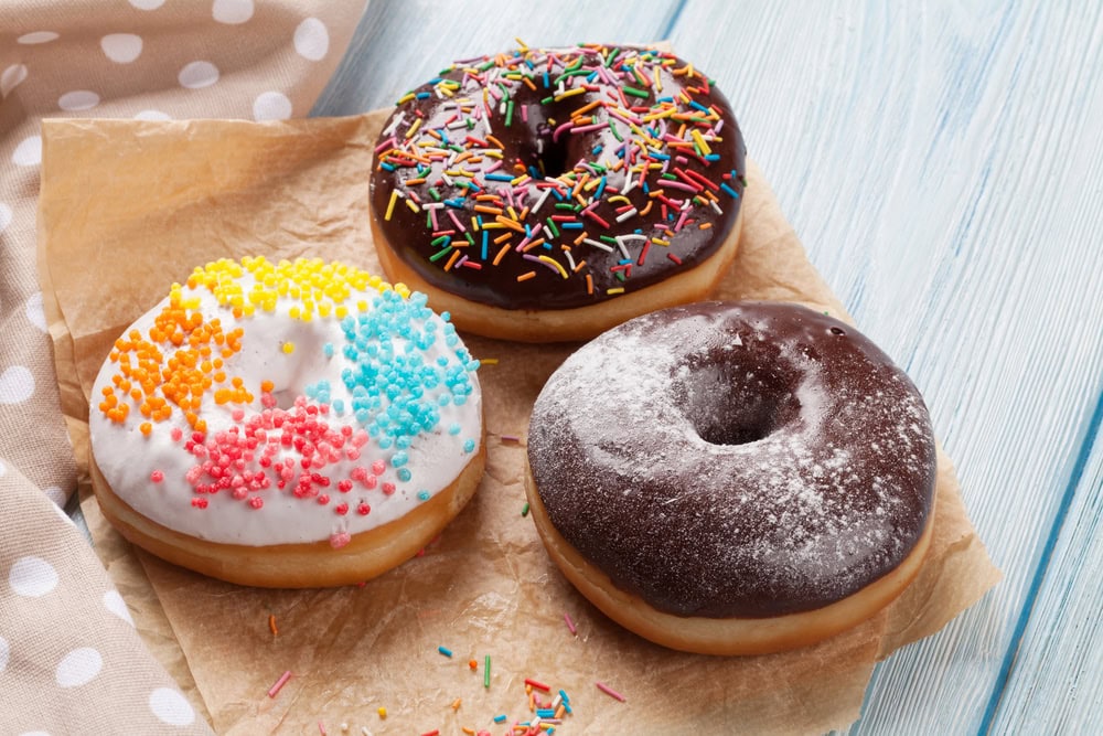 Three donuts on parchment paper: one with white icing and colorful sprinkles, one with chocolate icing and rainbow sprinkles, and one with chocolate icing and powdered sugar.