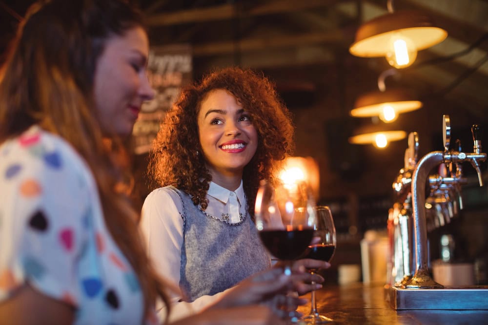 Two women sit at a bar holding glasses of red wine, talking and smiling. Beer taps and warm lighting are visible in the background.