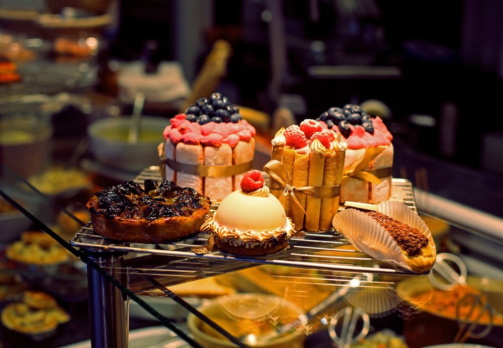 A display of assorted pastries and cakes, including fruit-topped tarts, mousse desserts, and layered cakes, arranged on a metal rack in a bakery setting.