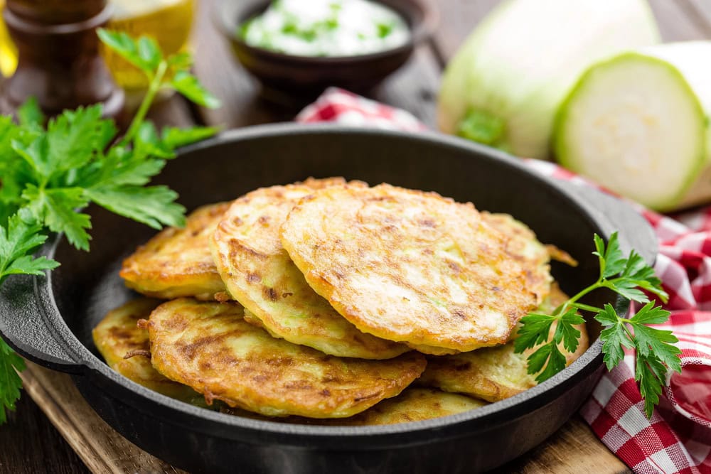 A black skillet holds several golden-brown zucchini fritters, garnished with parsley. A red checkered cloth, fresh zucchini, and a bowl of dip are in the background.