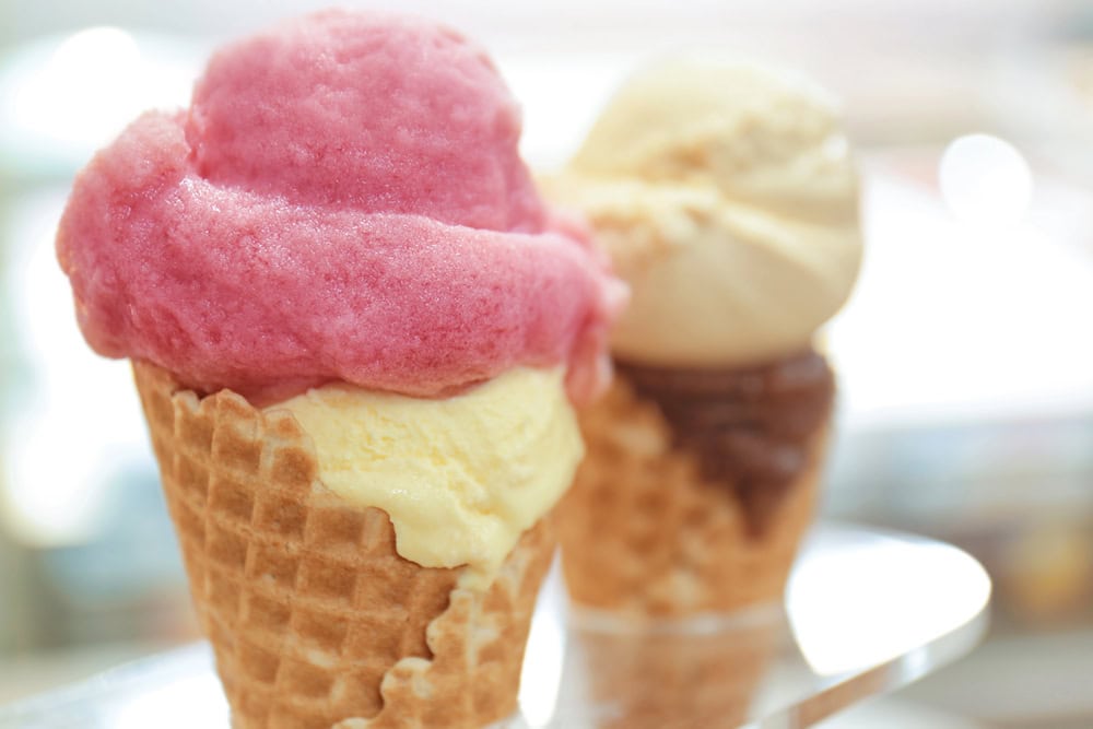 Two waffle cones with scoops of ice cream; one cone has pink and yellow ice cream, the other has brown and beige ice cream, both out of focus in the background.