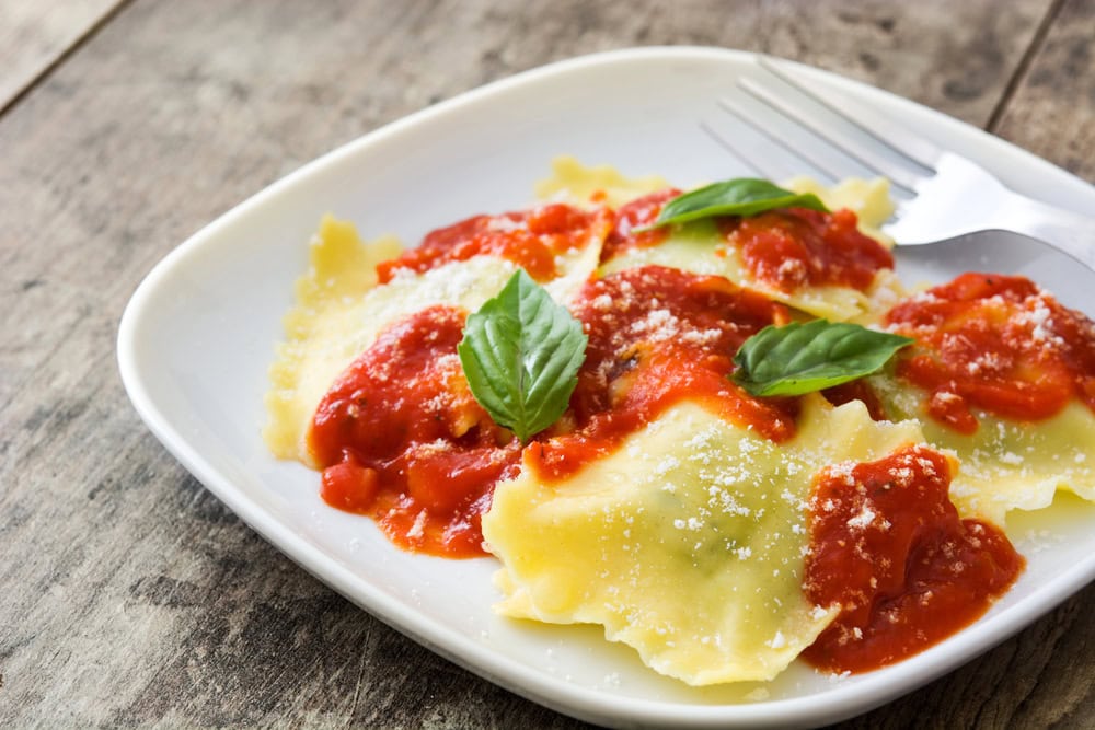 A plate of ravioli topped with tomato sauce, grated cheese, and fresh basil leaves, with a fork resting on the dish.