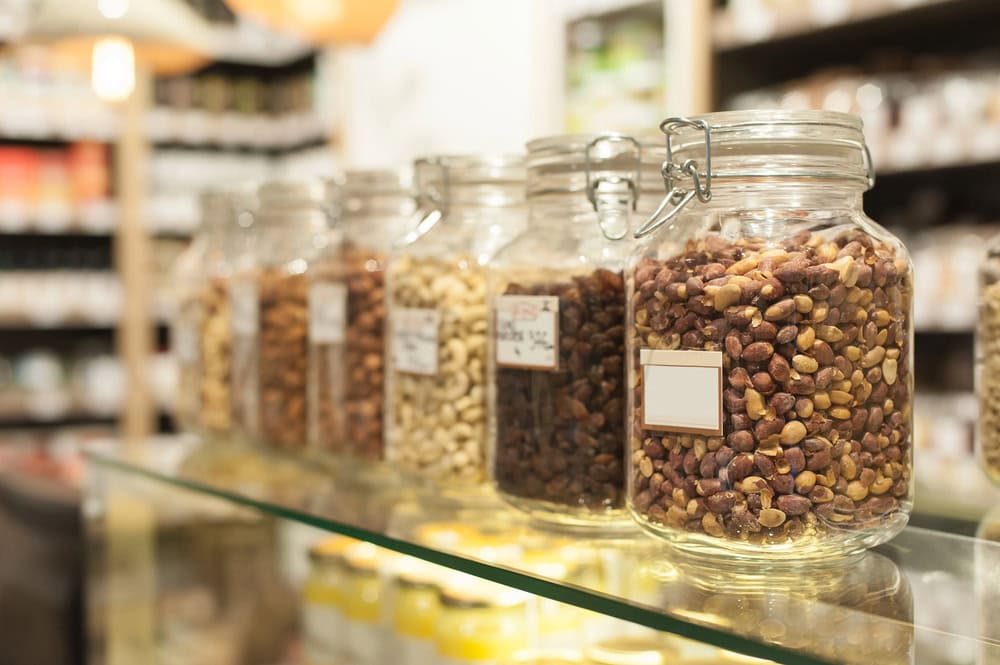 Five glass jars filled with various dried nuts and seeds are lined up on a glass shelf in a store, each with a blank label.
