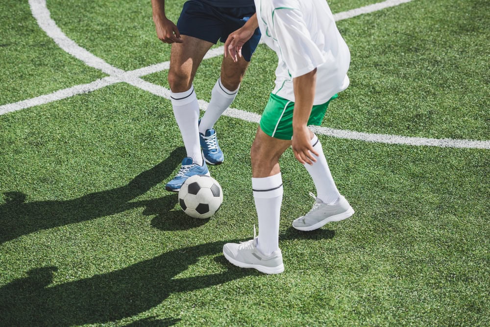Two soccer players, one in green shorts and the other in navy shorts, compete for the ball on a soccer field.