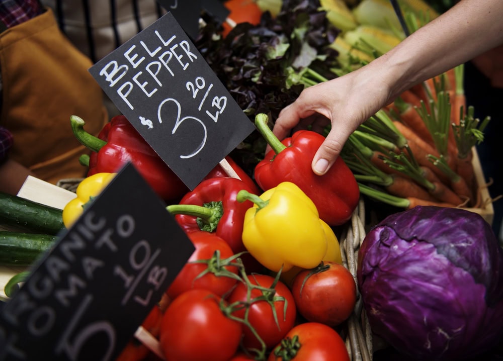 A person picks up a red bell pepper from a market stand displaying bell peppers, tomatoes, cucumbers, carrots, and cabbage with handwritten price signs.