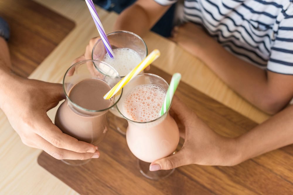 Three people clink glasses of milkshakes with colorful straws over a wooden table, viewed from above.