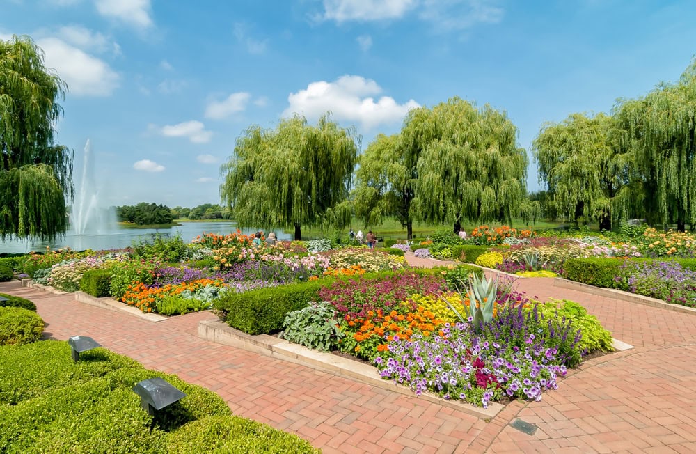 A landscaped garden with colorful flower beds, trimmed hedges, brick pathways, willow trees, and a fountain in the background under a blue sky.