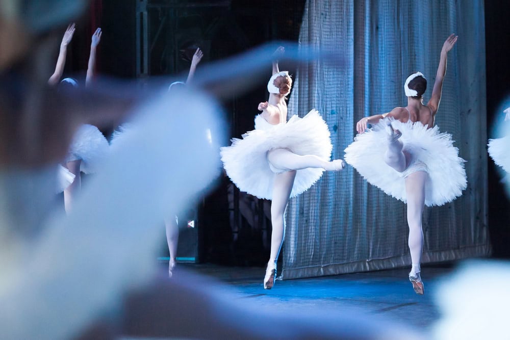 Ballet dancers in white tutus perform on stage, captured mid-leap with arms raised and one leg extended behind them.