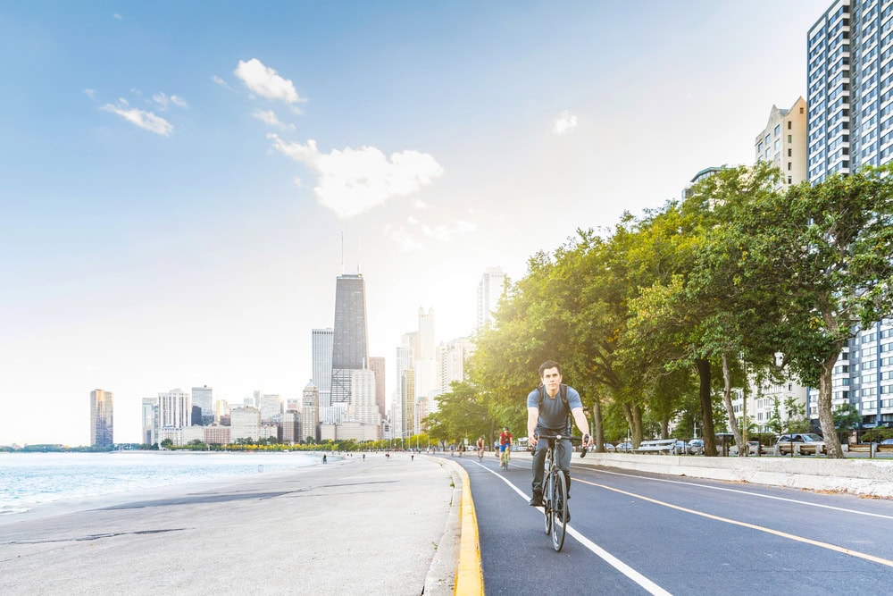 A man rides a bicycle on a lakeside path with tall city buildings and green trees in the background under a bright sky.