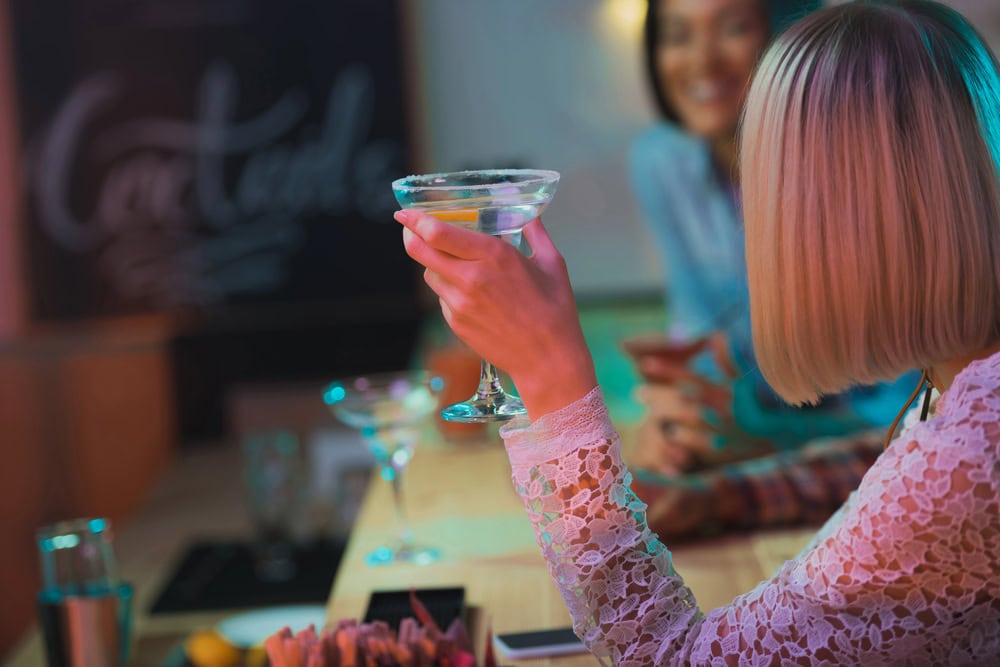 A woman with blonde hair holds a cocktail glass at a bar, sitting with another woman. Both are smiling and engaged in conversation.