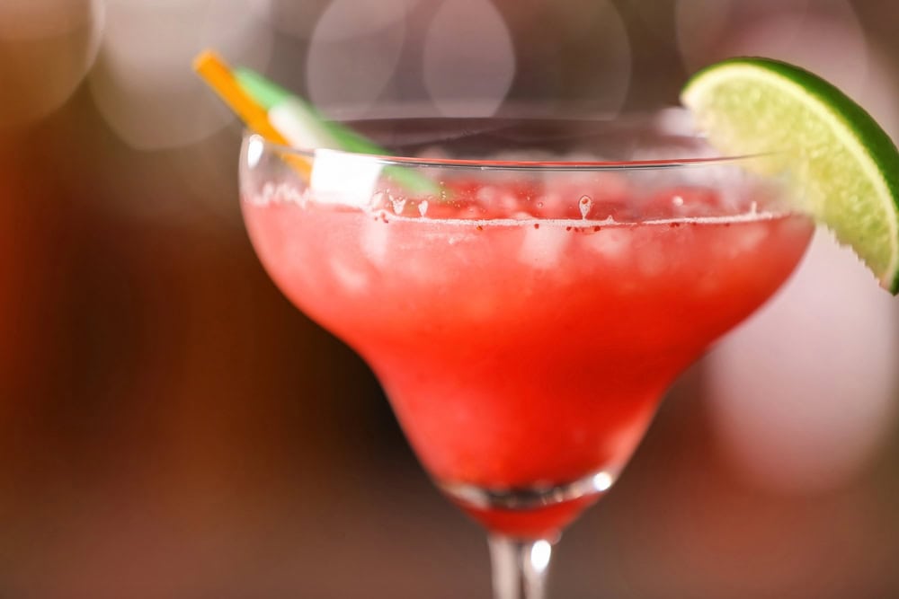 A close-up of a red frozen cocktail served in a margarita glass with crushed ice, garnished with a lime wedge and a straw.