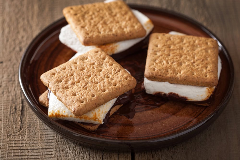 Three s'mores made with graham crackers, toasted marshmallows, and melted chocolate are arranged on a brown ceramic plate on a wooden surface.
