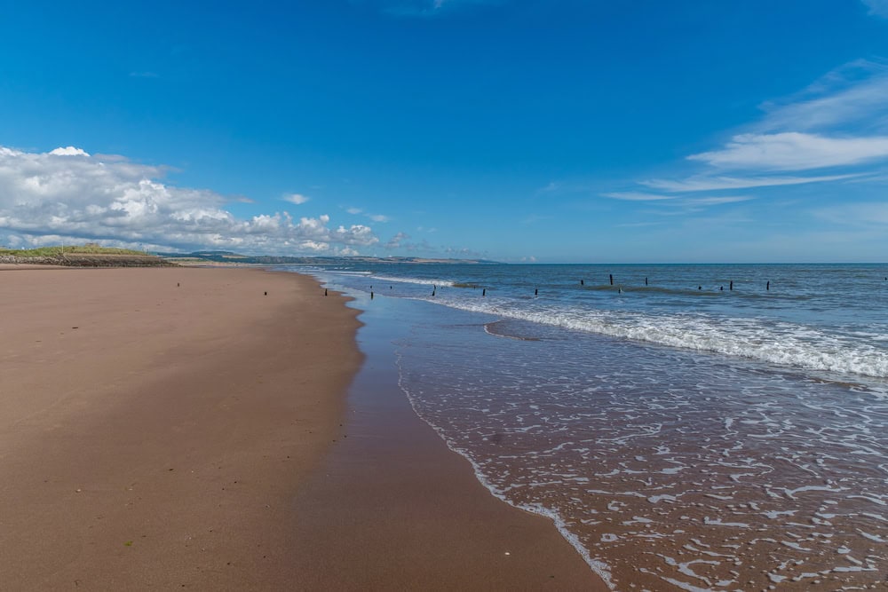 Wide sandy beach with gentle waves under a bright blue sky with scattered clouds; wooden posts visible near the shoreline.