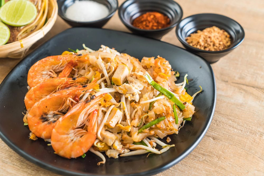 A plate of shrimp pad thai with tofu, bean sprouts, and green onions, served with three small bowls of condiments in the background.