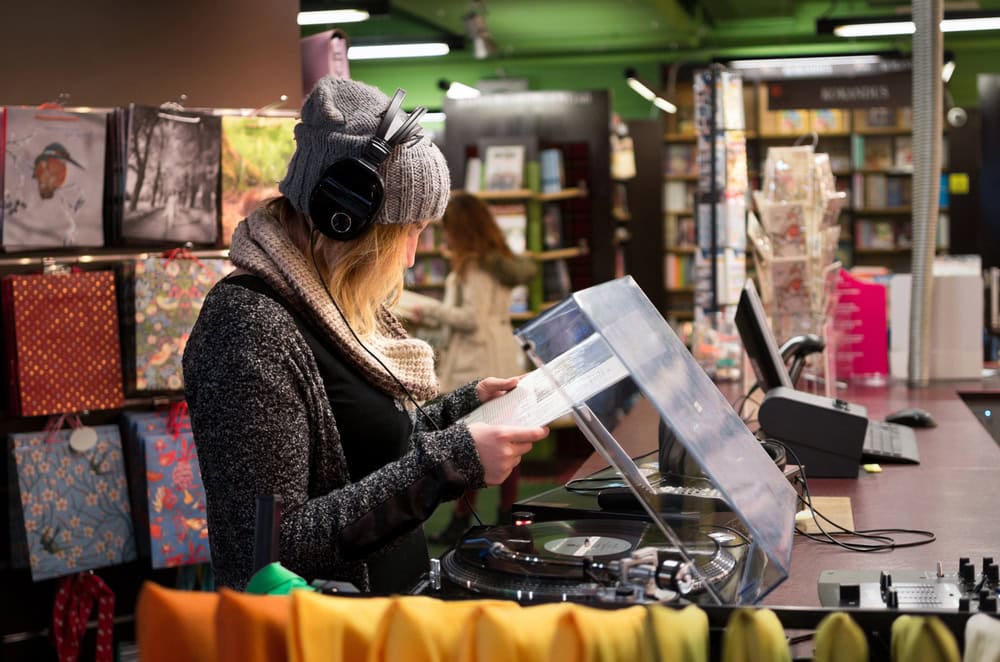 A person wearing headphones stands at a record player, examining a vinyl record in a store with gift bags, books, and cards on display.