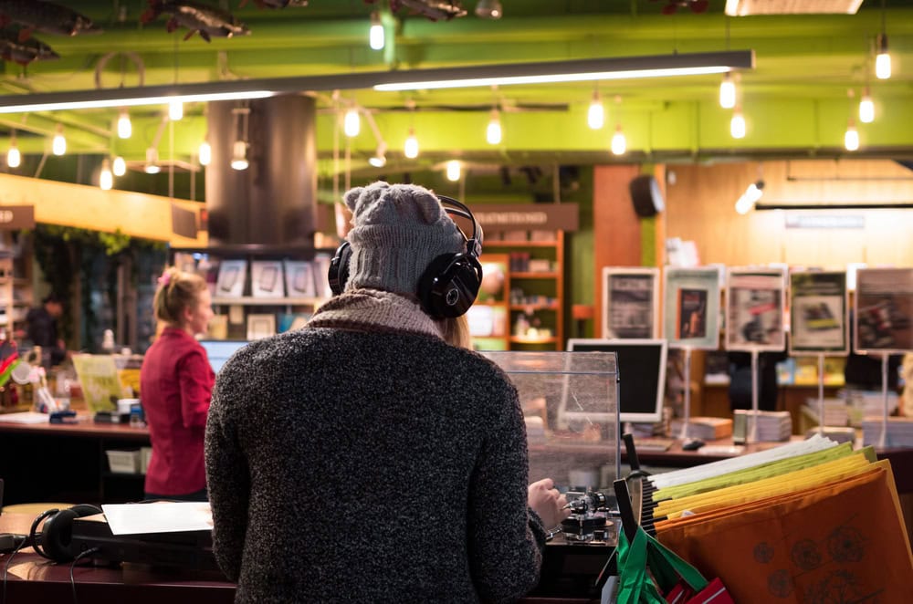 Person wearing headphones and a beanie stands behind a counter with equipment; another person in the background, surrounded by shelves and papers in a brightly lit indoor space.