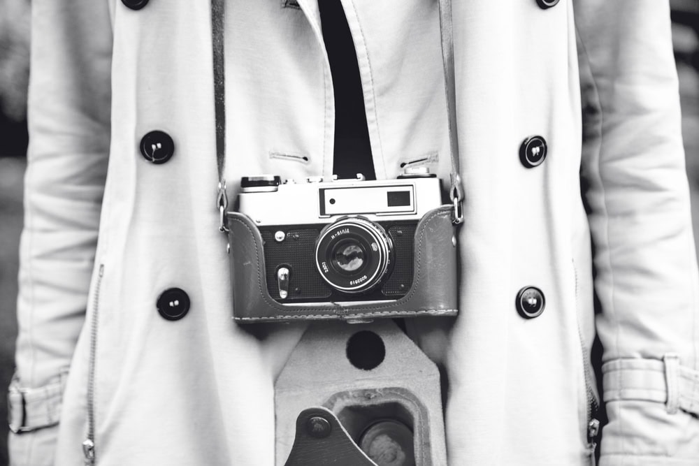 A person wearing a light-colored coat has a vintage film camera hanging from their neck by a strap. The image is in black and white.