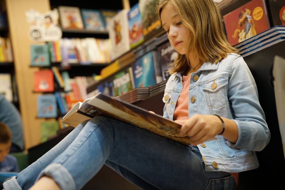 A girl in a denim jacket and jeans sits on the floor in a bookstore, reading a large book with shelves of colorful books in the background.