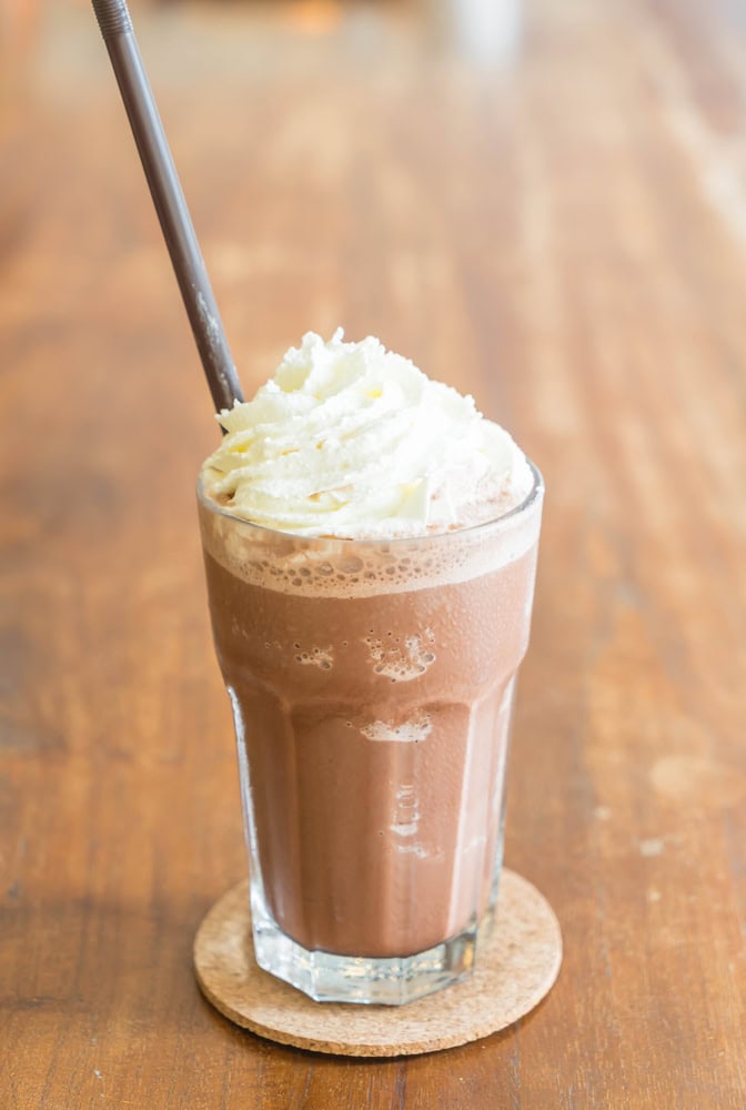 A glass of iced chocolate topped with whipped cream and served with a straw, placed on a cork coaster on a wooden table.