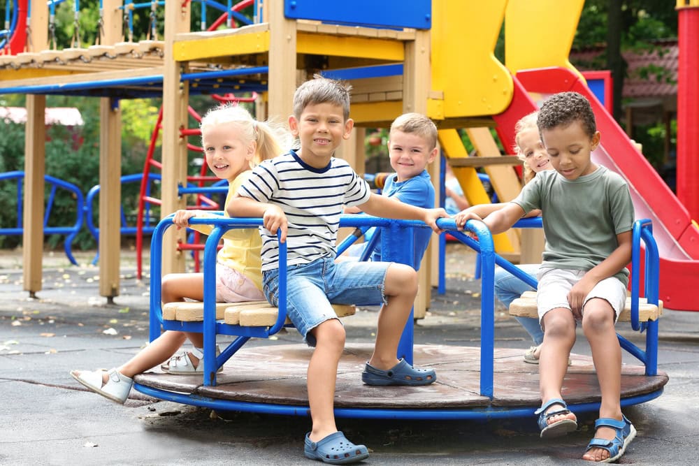 Four children sit on a blue and yellow merry-go-round at a playground, with slides and climbing equipment visible in the background.