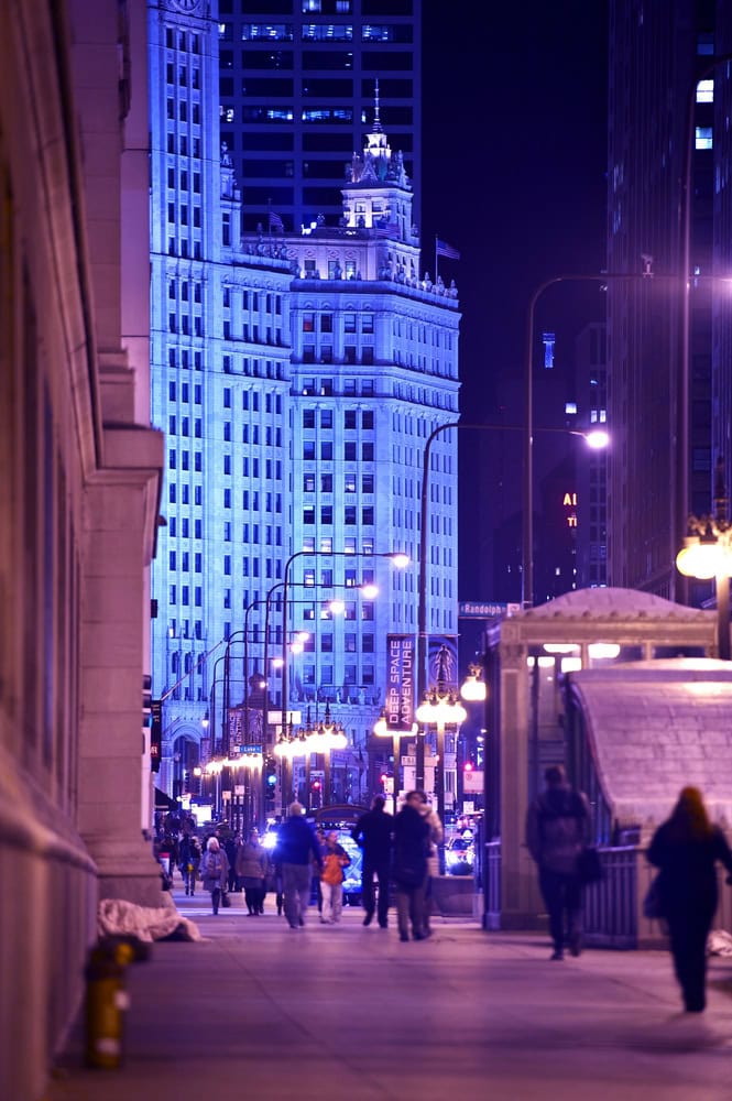 People walk along a city sidewalk at night, with tall buildings illuminated in blue light in the background.