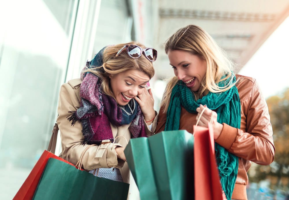 Two women wearing scarves and jackets look excitedly into shopping bags while standing outdoors, each holding several bags.