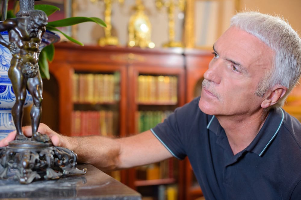 A man with gray hair examines a decorative metal figurine closely, with bookshelves and ornate objects in the background.