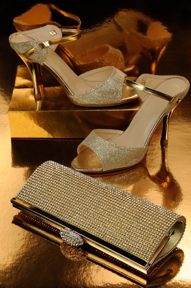 Gold high-heeled sandals and a matching rhinestone-studded clutch bag displayed on a reflective gold surface.