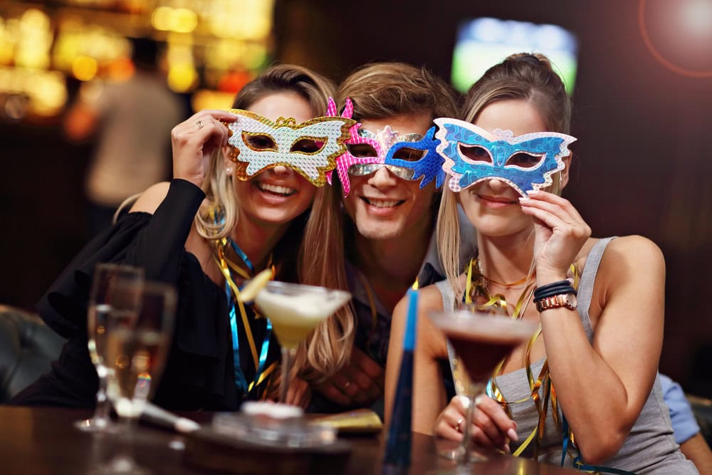 Three people at a bar wearing decorative masquerade masks, smiling at the camera. Cocktails and drinks are on the table in front of them.