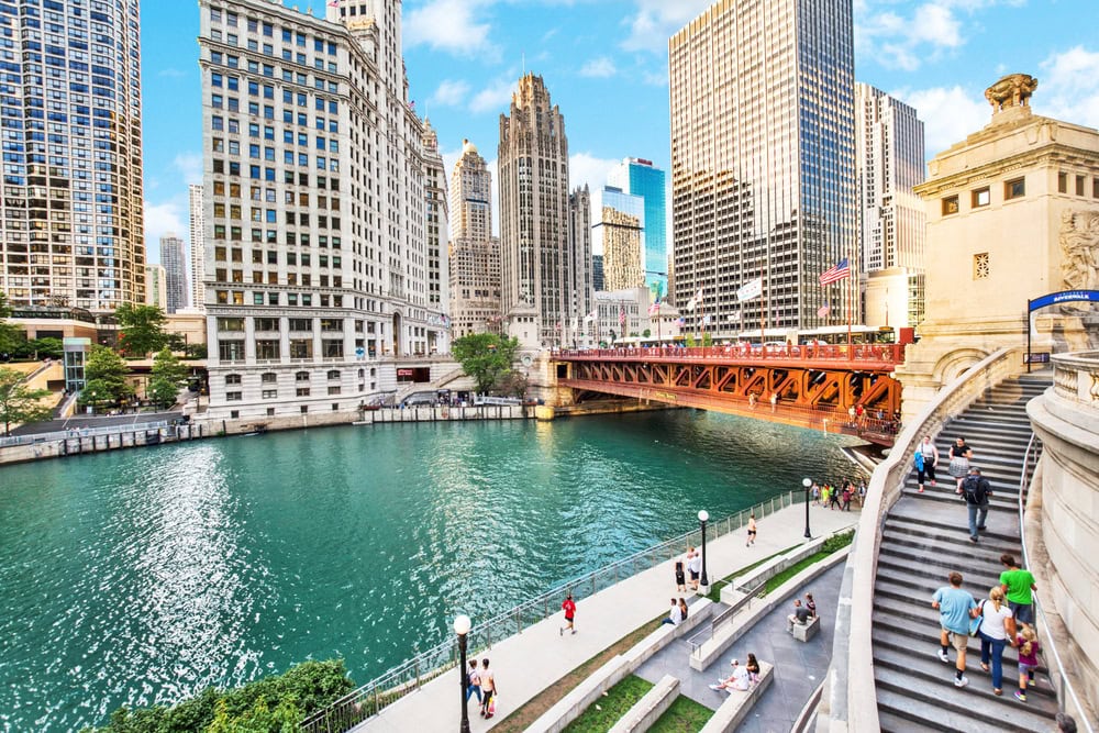 A view of the Chicago River with people walking along the riverside, surrounded by tall buildings and a red bridge under a partly cloudy sky.