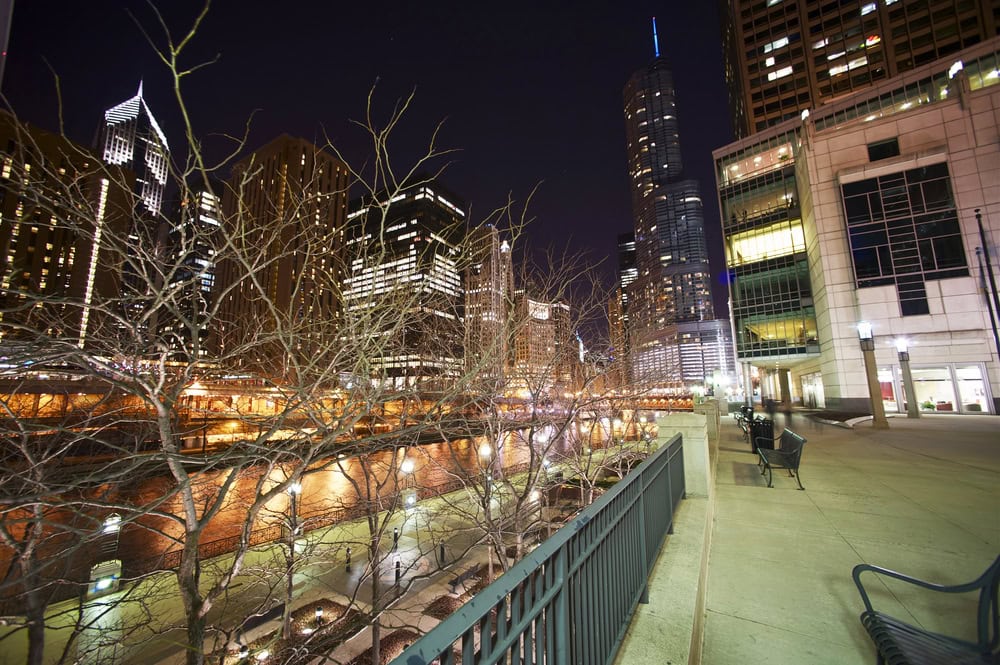 Cityscape at night with illuminated buildings, leafless trees, and empty benches along a riverside walkway. Skyscrapers are visible in the background.