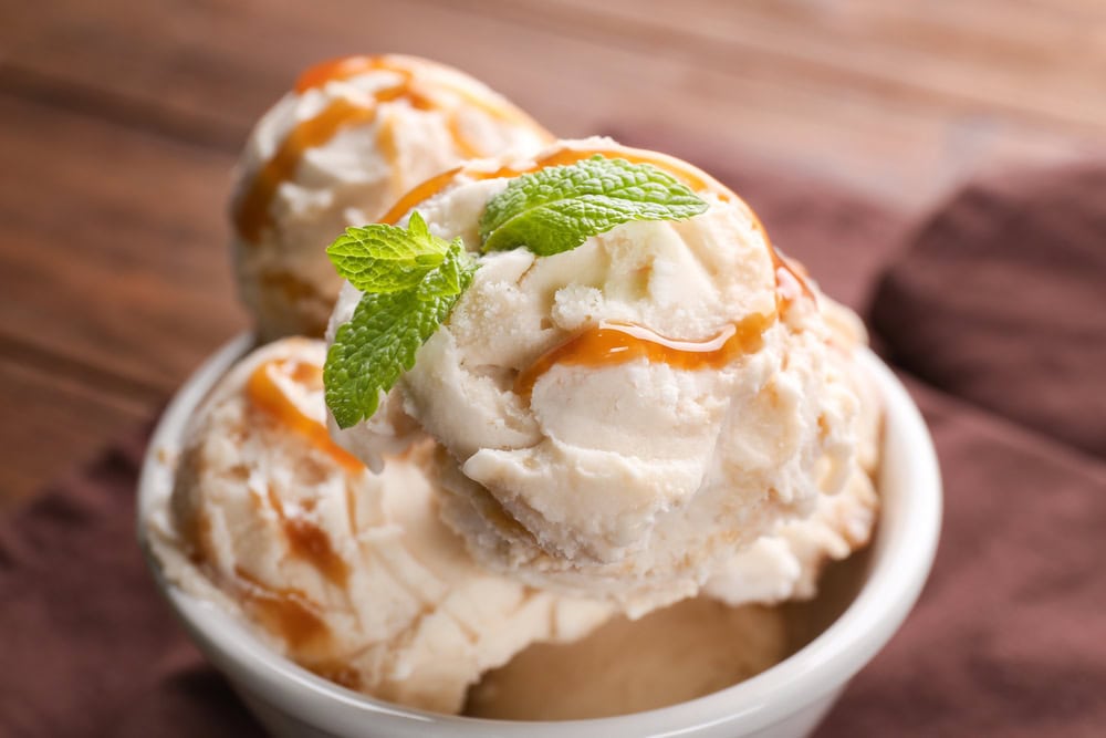 Three scoops of vanilla ice cream in a white bowl, topped with caramel sauce and a sprig of fresh mint, placed on a brown cloth.