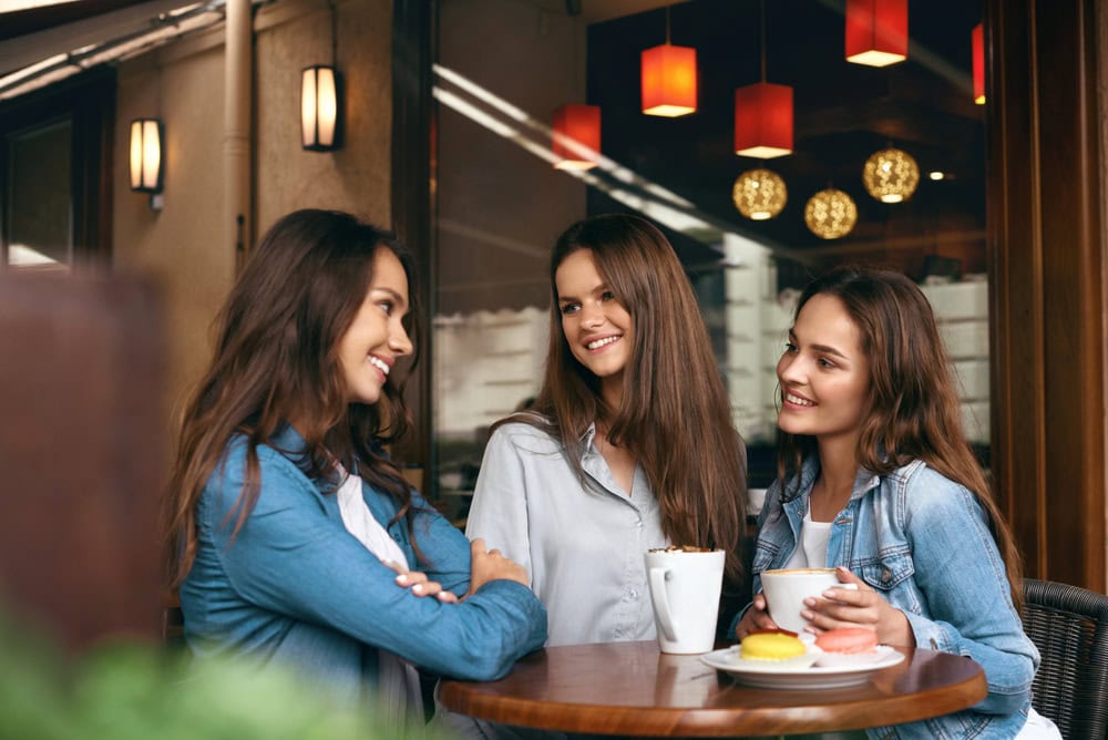 Three young women sit at an outdoor café table, smiling and talking, with coffee cups and a dessert in front of them.
