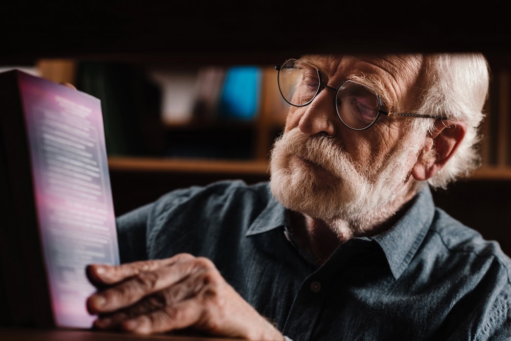 An elderly man with glasses and a white beard reads the back cover of a book while standing in front of a bookshelf.