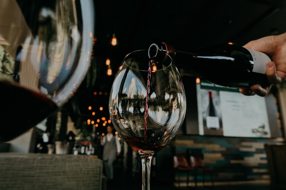 A person pours red wine into a glass in a dimly lit restaurant with hanging lights and a blurred background.