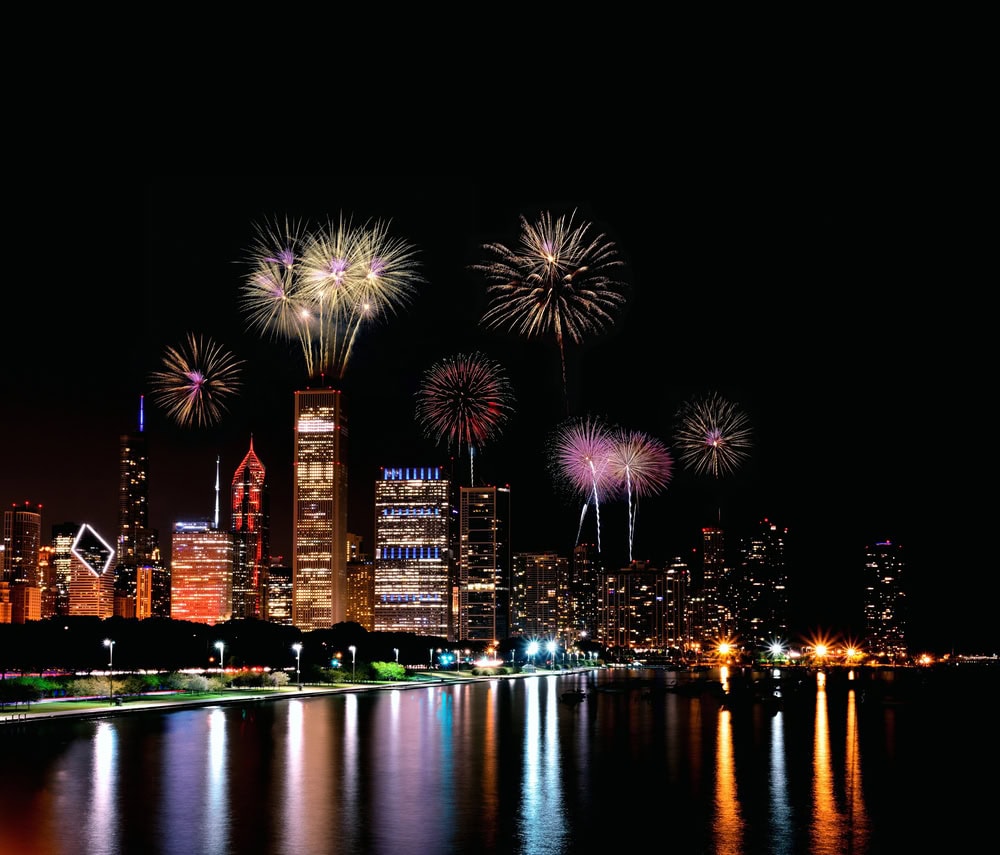 Fireworks illuminate the night sky above a city skyline, reflecting on the water below. Tall buildings are lit up against the dark background.