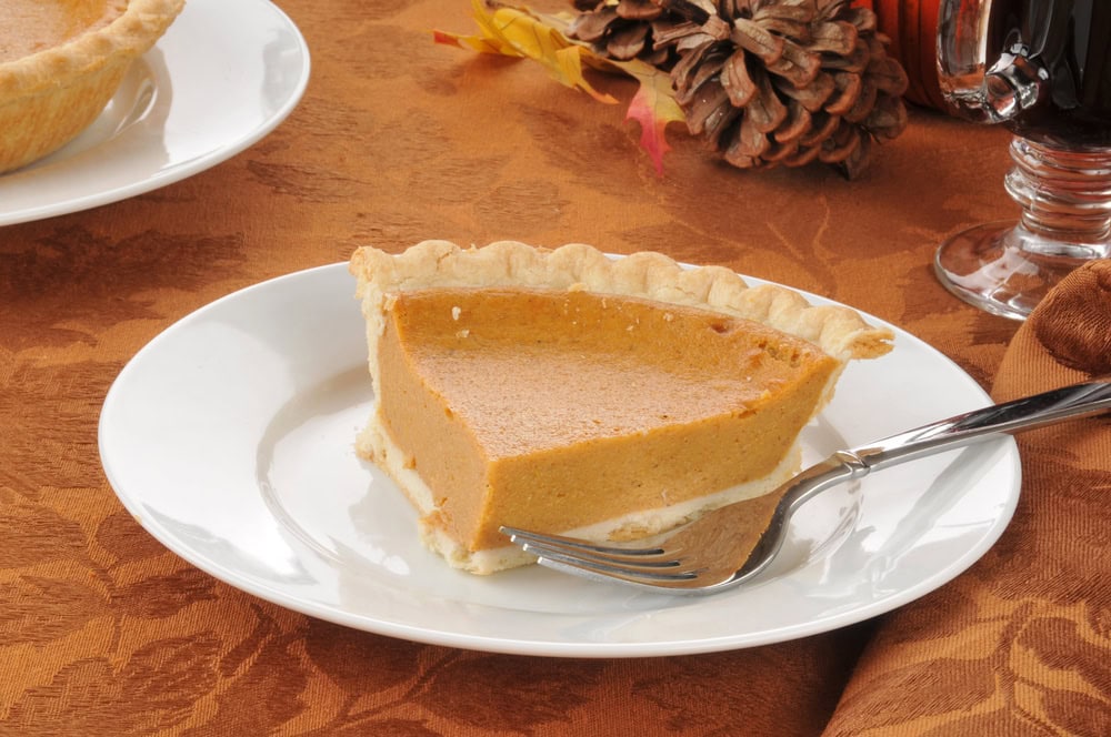 A slice of pumpkin pie on a white plate with a fork, set on a brown tablecloth with autumn decor in the background.