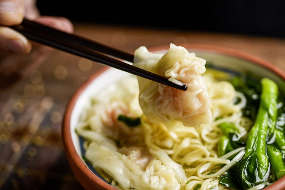 A close-up of chopsticks holding a dumpling over a bowl of noodles with green vegetables.