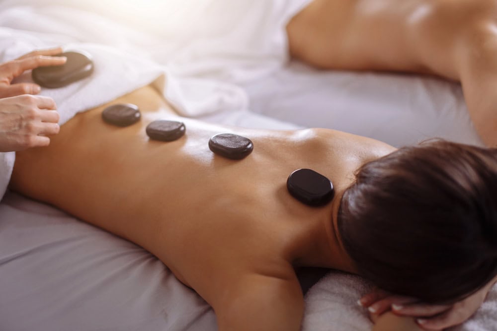 Two people receiving a hot stone massage, lying face down on massage tables with heated stones placed along their spines.