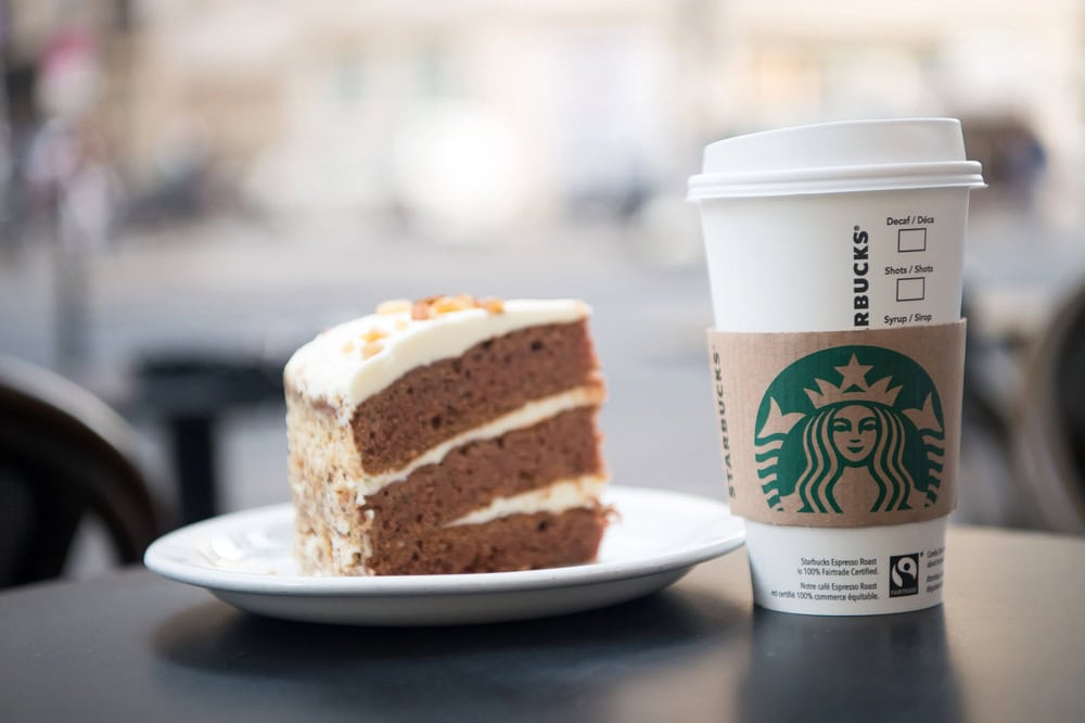 A slice of layered cake on a white plate next to a Starbucks paper cup with a lid and sleeve, on a dark table.