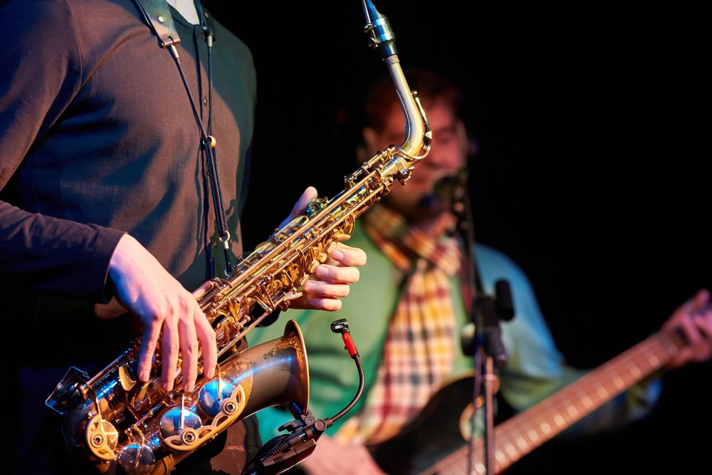 A person plays a saxophone on stage while another musician in the background plays a guitar and sings into a microphone.