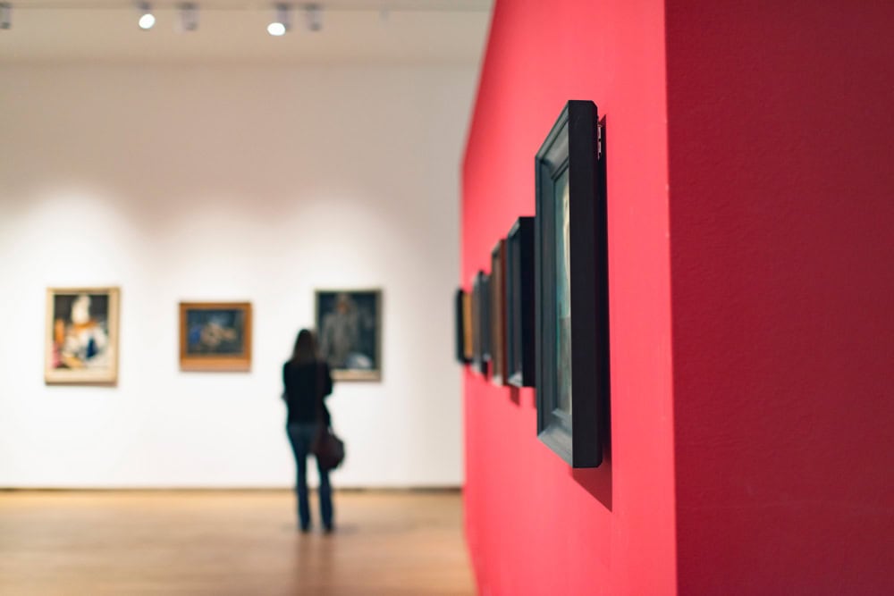 A person stands in an art gallery, looking at paintings on a white wall; framed artworks are also displayed on a bright red wall in the foreground.