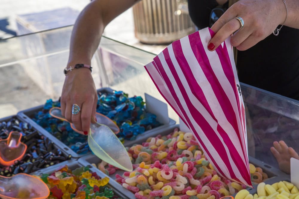 A person scoops assorted gummy candies into a pink and white striped bag at an outdoor candy stand.