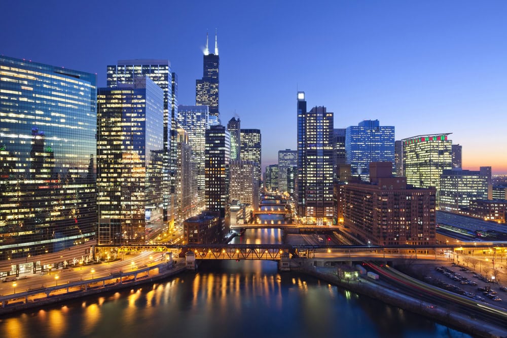 City skyline at dusk with illuminated office buildings, river, and bridges; clear blue sky in the background.