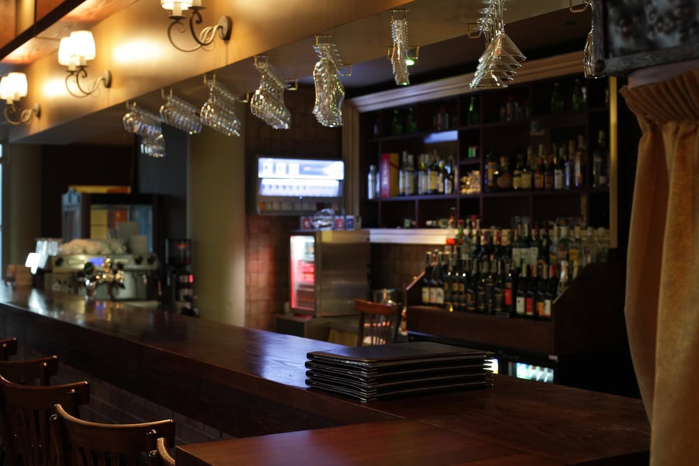 Empty bar counter with stacked menus in the foreground, overhead glasses, and shelves of bottles and a coffee machine in the background.