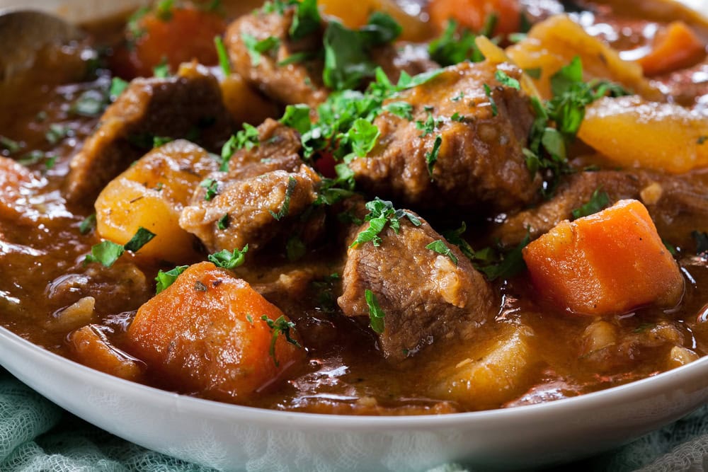 A close-up of a hearty beef stew with large chunks of beef, carrots, potatoes, and garnished with chopped parsley.